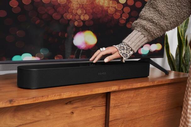 Schwarze Sonos Beam Soundbar unter TV-Gerät (Foto: Sonos) Frau bedient Touch-Feld der Sonos Beam (2. Gen) Soundbar